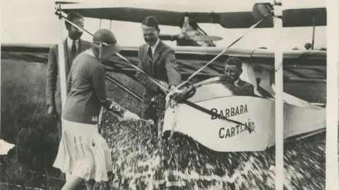 Cartland Promotions Black and white photo of Barbara seen from behind, in front of the glider titled Barbara Cartland. She appears to be breaking a bottle of fizz onto the plane - it is not clear. She wears a pale knee length skirt, dark cardigan and flapper-style hat. A man sits in the pilot's seat and two other men stand next to the plane.