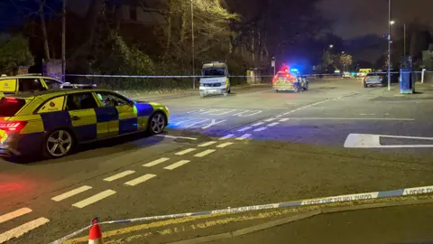 Eleanor Moritz/BBC The scene of a fatal collision on a road at night, the area is cordoned off with police tape and there are several police cars at the scene with flashing lights. 
