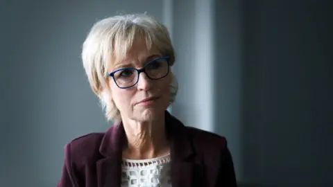 Sally Magnusson sits successful  a darkened country   wearing blue-rimmed glasses, a achromatic  apical  and a burgundy blazer