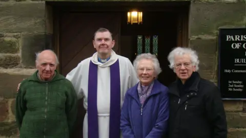 The Friends of the Church of Our Lady The Reverend David Atkinson wearing his vestments. He is standing with two men and a woman from the parish outside of the church.