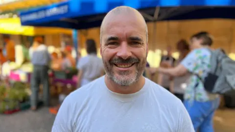 A man with a shaved head and short beard stands outdoors in front of market stalls. He's wearing a plain white T-shirt and faces the camera, while canopies, plants and several people are visible in the background.
