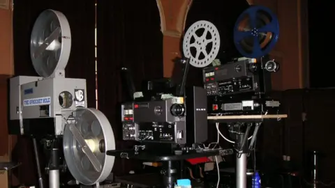 John Clancy Multiple old projectors side by side, some with spools of film attached. One has a WB Warner Bros logo.