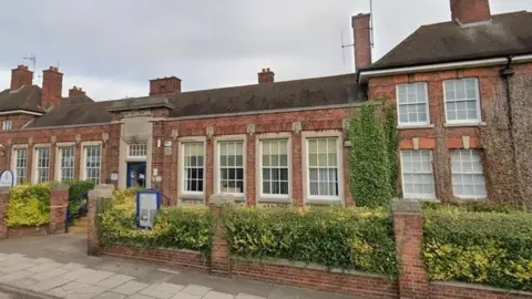 A red-bricked building with white framed Georgian bar-style windows. There is a low wall with pillars at the front with hedging between the pillars and sign near the entrance.