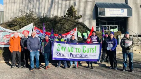 A group of about a dozen people holding protest banners outside an Aberdeenshire Council building.