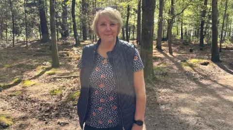 BBC A woman with shoulder length light hair is standing in a forest landscape with the trunks of trees behind her. It is a sunny day. She is wearing a blue patterned top with a navy blue body warmer and a black watch. 
