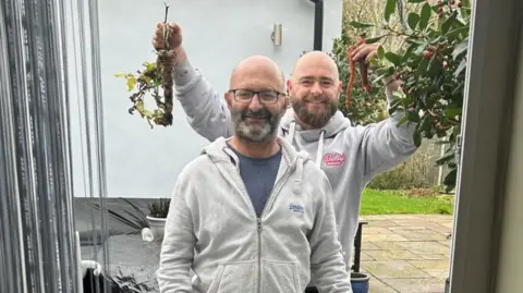 Brain Tumour Research Two men can be seen smiling, with the one at the back holding up vegetables in both hands.