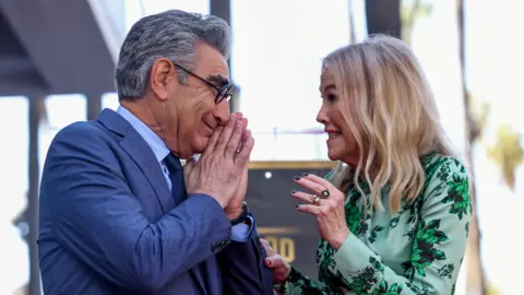 Reuters Eugene Levy and Catheirne O'Hara face each other and smile. Levy is an elderly man with short grey hair, wearing black circular glasses and holding his hands to his mouth. He wears a blue jacket, light blue shirt and a dark tie. O'Hara has short blonde hair and wears a floral-patterned green dress with long sleeves.