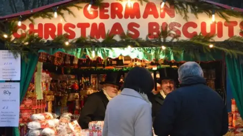 A biscuit stall at Lincoln Christmas Market showing two servers dressed in Victorian costume, including top hats. A sign, in large red letters, reads "German Christmas biscuits".