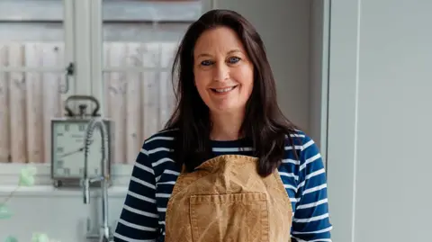 Kiersten is wearing a horizontally, dark blue and white striped, long sleeved top.
She is also wearing a light brown apron and is standing by a table in a mainly white coloured kitchen.