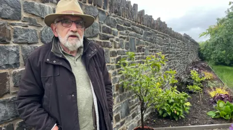 John Watkeys standing against his new stone garden wall which is about head height. He is wearing a fawn-coloured hat and glasses with a black coat and has a white beard. A flower bed can be seen to the right of the wall and grass to the right of that. 