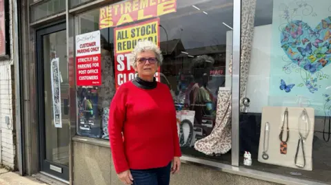 Pauline Crawley A woman with short curly white hair and wearing a red jumper stands in front of a shop window which has a large red sign saying, 'Retirement Sale'.