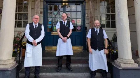 Empire Hotel Jean Paul, Nelson and David all wearing white shirts, black ties, black waistcoats  and white aprons standing in front of the hotel.