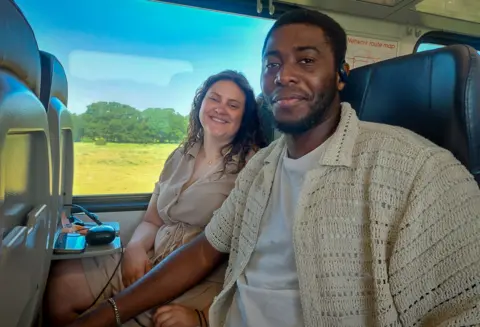 A man with short dark hair wearing a beige unbuttoned shirt and white T-shirt sat on a train next to a woman with long dark hair wearing a beige dress