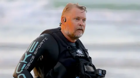 A man wearing a wetsuit and an earpiece stands in front of the sea on a beach.