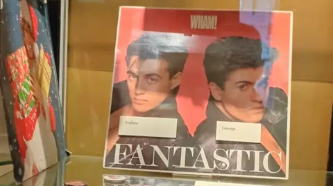 Bushey Museum and Art Gallery A glass display cabinet dedicated to the pop duo Wham!, containing a variety of memorabilia and publications arranged across several glass shelves. On the central shelf is a large Wham! “Fantastic” album cover, featuring two portrait photographs and the word “FANTASTIC” in large lettering across the bottom.