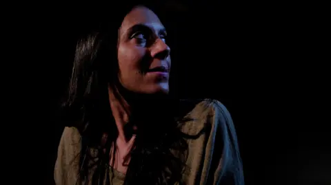 Laurence Cawley/BBC A side profile of Nadine Buzzard-Quashie who has long black hair and wears a dark grey top in front of a black background