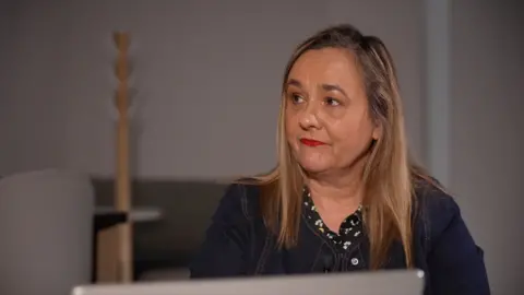 Dr Emma Cunningham, a woman with dark blonde hair and a navy blue jacket. She is looking off camera and is sitting in front of a laptop screen