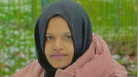 Family handout Kulsuma Akter - a young woman wearing a black headscarf and salmon-coloured coat. She is pictured in what appears to be a park.