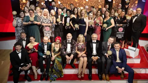 VisitEngland A large group of men and women in either suits or stylish dresses stand on the stage at the Visit England awards, with spotlights overhead