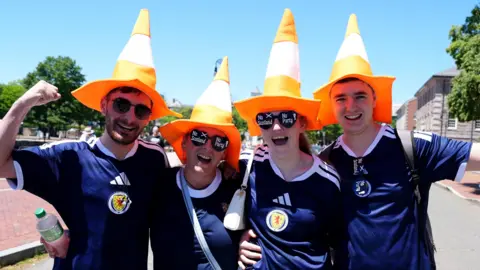 Four Scotland football fans wearing blue Scotland football shirts and orange traffic cone hats. They are cheering towards the camera.