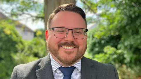 Lewis Adams/BBC Daniel Cowan is wearing glasses, a grey suit jacket, blue and white striped shirt and navy tie. He has short brown hair and is smiling in front of bushes.