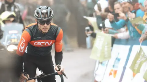 Andrew Colele é retratado por Andrew Feder em sua bicicleta, ele usa capacete e óculos escuros com equipamento de ciclismo vermelho e preto. 