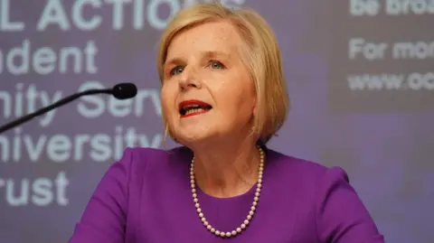 PA Media Donna Ockenden speaks into a microphone. She has blonde hair in a bob, is dressed in purple and is wearing pearls