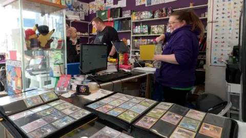 A woman wearing a purple hoody and with brown hair in a ponytail is standing behind a till, on one side is binders full of Pokémon cards and on the other is the till with customers behind it.