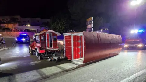 A small red tourist train, with its passenger wagon lying sideways on the street. Some emergency service vehicles can be seen near it.