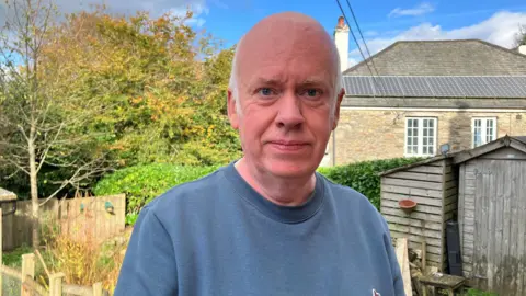 A man with a bald head and white side burns, wearing a blue sweatshirt. He's standing in a garden where the trees are showing their autumnal colours and there is a stone walled building with solar panels on the roof.