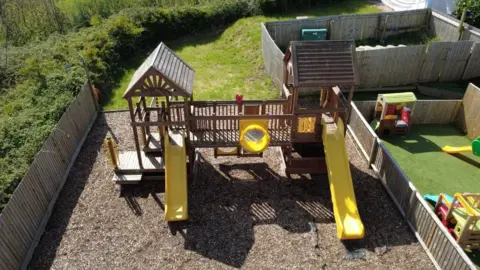 Gary Bulmer An outdoor play area, fenced in by wood panels, featuring two yellow slides, a bridge, a window in the centre, and monkey bars. The ground surrounding the play area is covered in bark, and the area is surrounded by greenery.