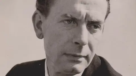 John Crowe A black and white photo shows a man looking to the bottom right of the image, not meeting our gaze. He is clean shaven with a slight frown on his forehead and wears a dark suit jacket over a lighter shirt.