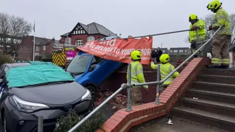 Nottinghamshire Fire and Rescue Service A blue car has gone over a wall and come to rest, at an angle, against another vehicle and beside a set of steps, while firefighters examine the scene