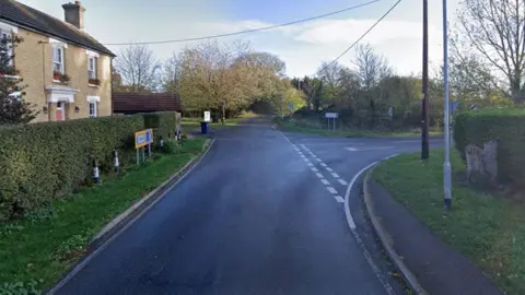 Google A view of the junction from Hilton Road into Connington. There is a house on the left side, behind a hedge, and a road sign to Connington and Fenstanton next to the junction. There are telegraph wires overhead and two posts on the right. Trees and fields are in the distance.