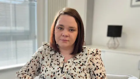 Nikella has shoulder length brown hair. She is sitting on a greychair in a cream-coloured living room. She is wearing a white top with a black and brown spot pattern. A cabinet with a lamp sitting on top are in the background.