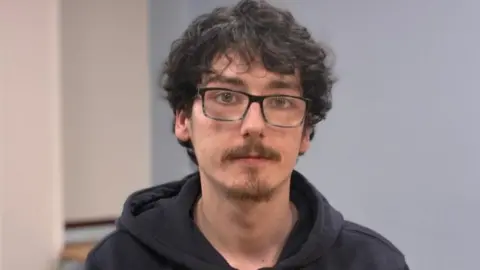Conner is a white male with messy black hair that hangs down to his eyes. He is wearing glasses and looking straight at the camera. His face is serious. He has a thin haired mustache and goatie. Conner is wearing a black hoody. 