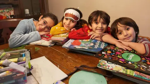 Left to right, Hajar, Amina, Zakaria and Ismaeel sit around a table with their arms folded and their chins resting on their hands, looking happy. They are surrounded by books.