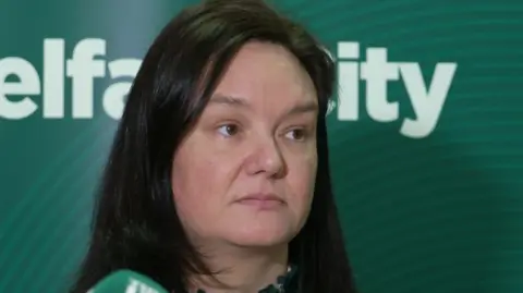 Detective Chief Inspector Gina Quinn has brown long hair and brown eyes. She is looking to the side with a microphone in vision and a green background with white writing that isn't readable. It says 'elfa... ity'