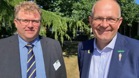 BBC/CARA THORPE Two men smile at the camera. The man on the left is wearing a grey blazer, a blue-and-white checked shirt and a blue and yellow tie. The man on the right is wearing a blue blazer and a shirt with thin blue-and-white stripes. Both are wearing National Farmers' Union badges.