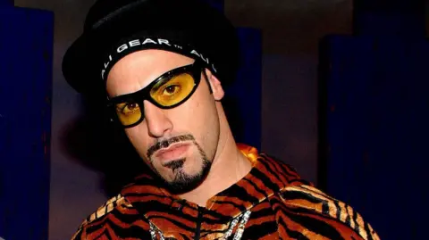 A man with a black goatee looks at the camera, wearing a tiger print jacket, thick gold chains around his collar, and black-rimmed yellow sunglasses. He is wearing a black hat, with a band across his forehead that reads "ALI GEAR TM".