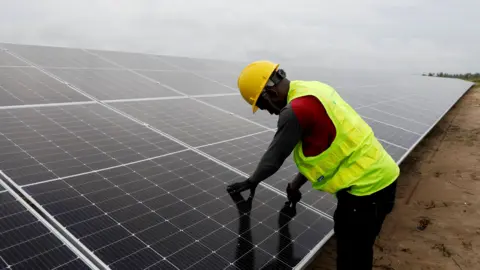 A man in a yellow high visibility jacket and yellow helmet can be seen working on a large solar installation.