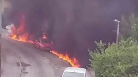 A strip of flames runs down a road. In the centre, you can se a small red car with a black cross on its front. Lots of black smoke blocks the view behind the road.