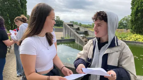 Jacob Redwood wearing a grey jumper with his hood up and a beige and blue jacket. He is standing beside a girl wearing a white t-shirt, and they are both holding they A-Level results and looking at each other