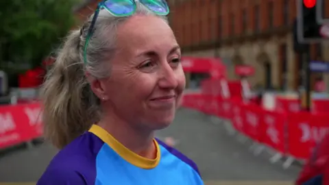 A woman with long grey hair and wearing a turquoise, indigo and yellow top and sunglasses resting on top of her head is smiling as she stands next to the route.