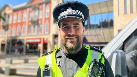 Shariqua Ahmed/BBC A man, wearing a green and black police officer uniform and a black hat, smiling for the camera.