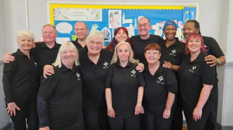 Hull Deaf Centre A group of 12 people stand shoulder to shoulder in two rows in a white-walled room and smile at the camera. They are made up of seven women and four men, all wearing black polo shirts with white logos. Behind the group is a blue and yellow picture board with hand-drawn pictures and artwork.