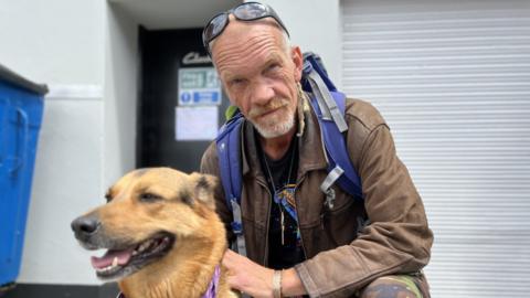 Martin looking at camera while wearing brown leather jacket and rucksack, holding a German Shepherd-type dog called Josie. Martin is crouching with his hand on the dog's collar in front of commerical building with large blue wheelie bin to his right