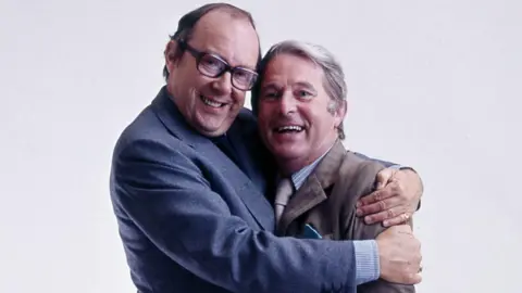 Close up of Eric Morecambe with his arms around Ernie Wise giving him a big hug. Both are smiling, Eric is wearing a blue suit, has receding dark hair and dark-framed glasses. Ernie is in a brown suit and has short white hair