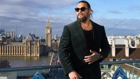 PA Media Jason Momoa standing on a balcony on the south side of the River Thames in London. Behind him are County Hall and, across the river, the Houses of Parliament. He is wearing a black suit jacket over a black t-shirt, with his left hand (sporting a giant green ring on his index finger) laid across his midriff. He has brown framed sunglasses and is looking slightly to his right. There are logos for the DC film Aquaman on the glass panels of the balcony.