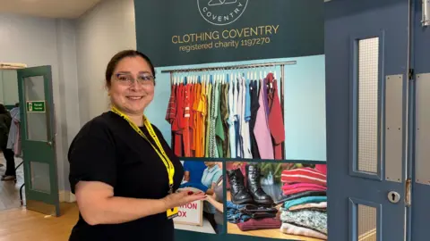 Seyedeh Naserinaki has dark brown hair tied back and wears glasses. She stands with a smile on her face and a yellow lanyard around her neck. She is pointing to a Clothing Coventry banner behind her.